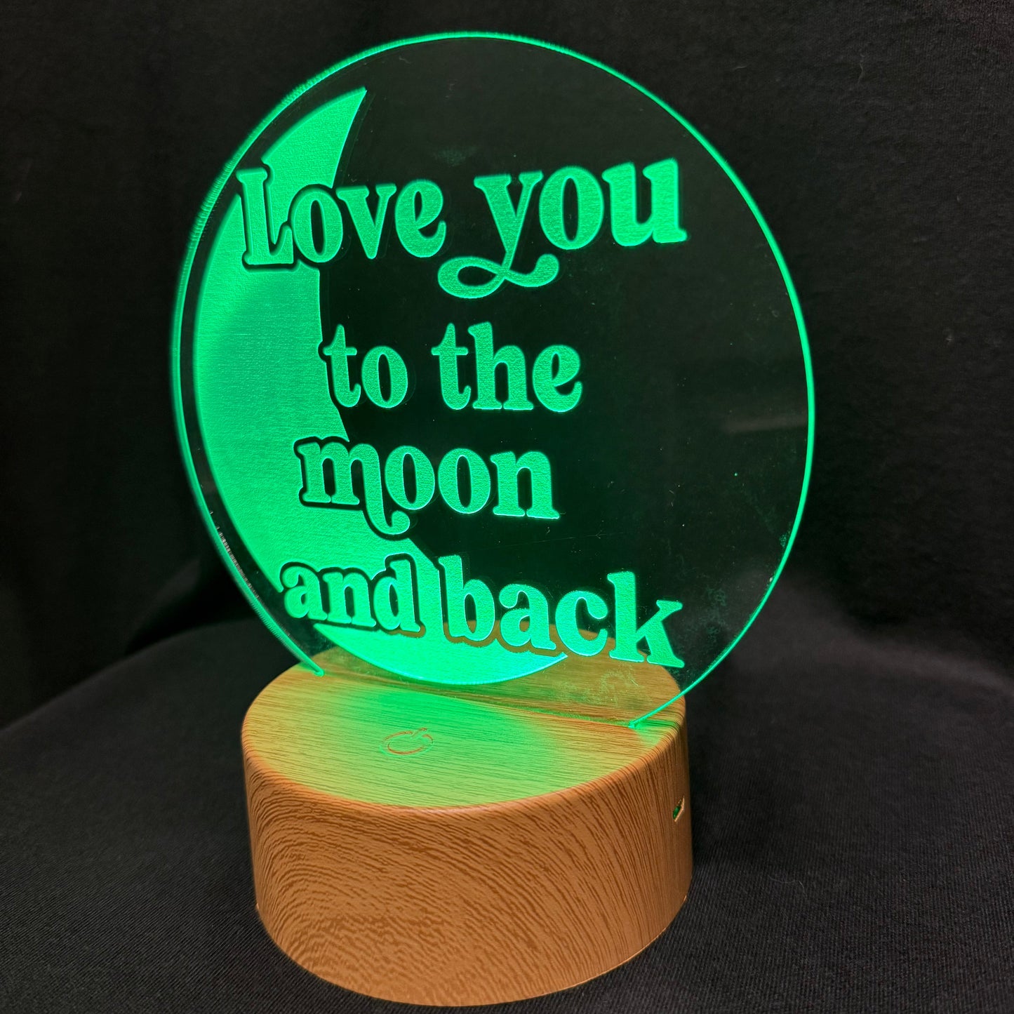 A circular acrylic LED night light with the phrase 'Love you to the moon and back' in glowing green text, mounted on a cylindrical wooden base against a black background.