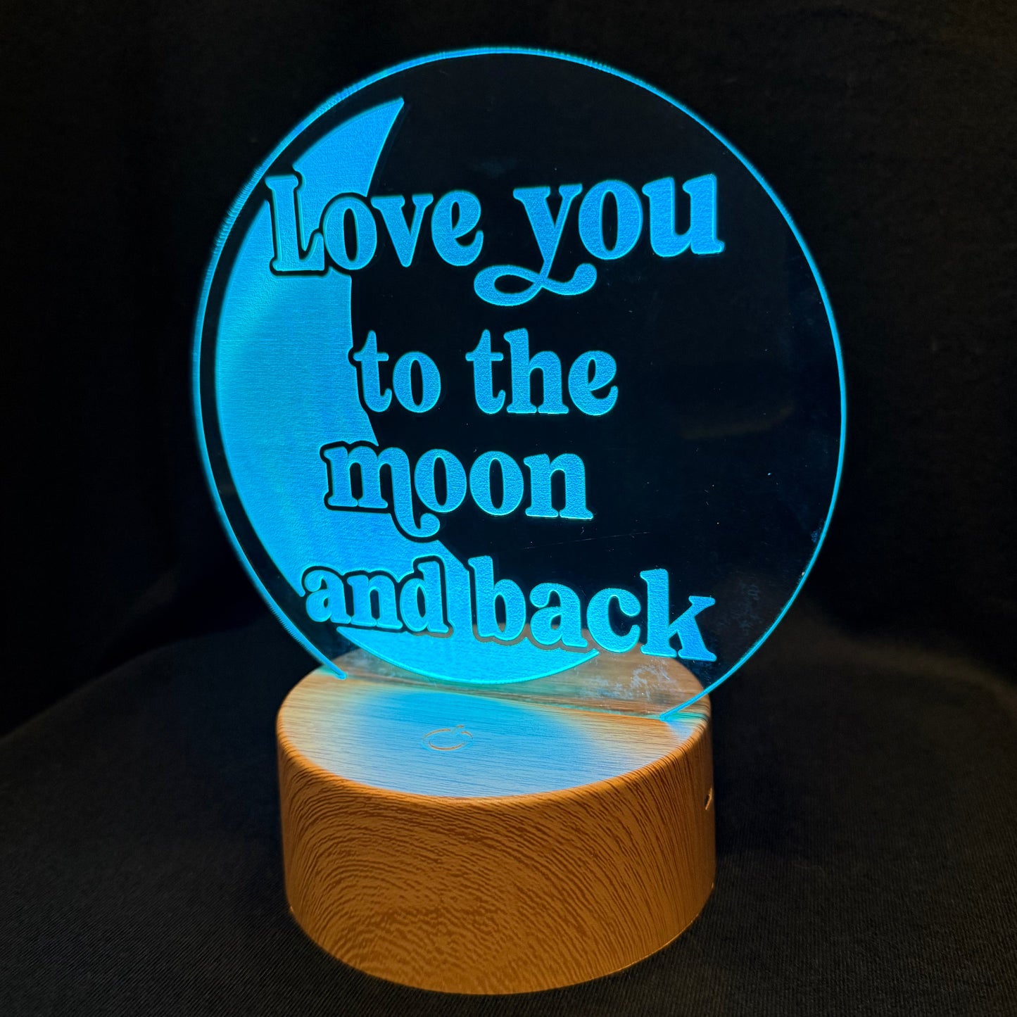 A circular acrylic LED night light with the phrase 'Love you to the moon and back' in blue lettering, mounted on a cylindrical wooden base and glowing against a dark background.