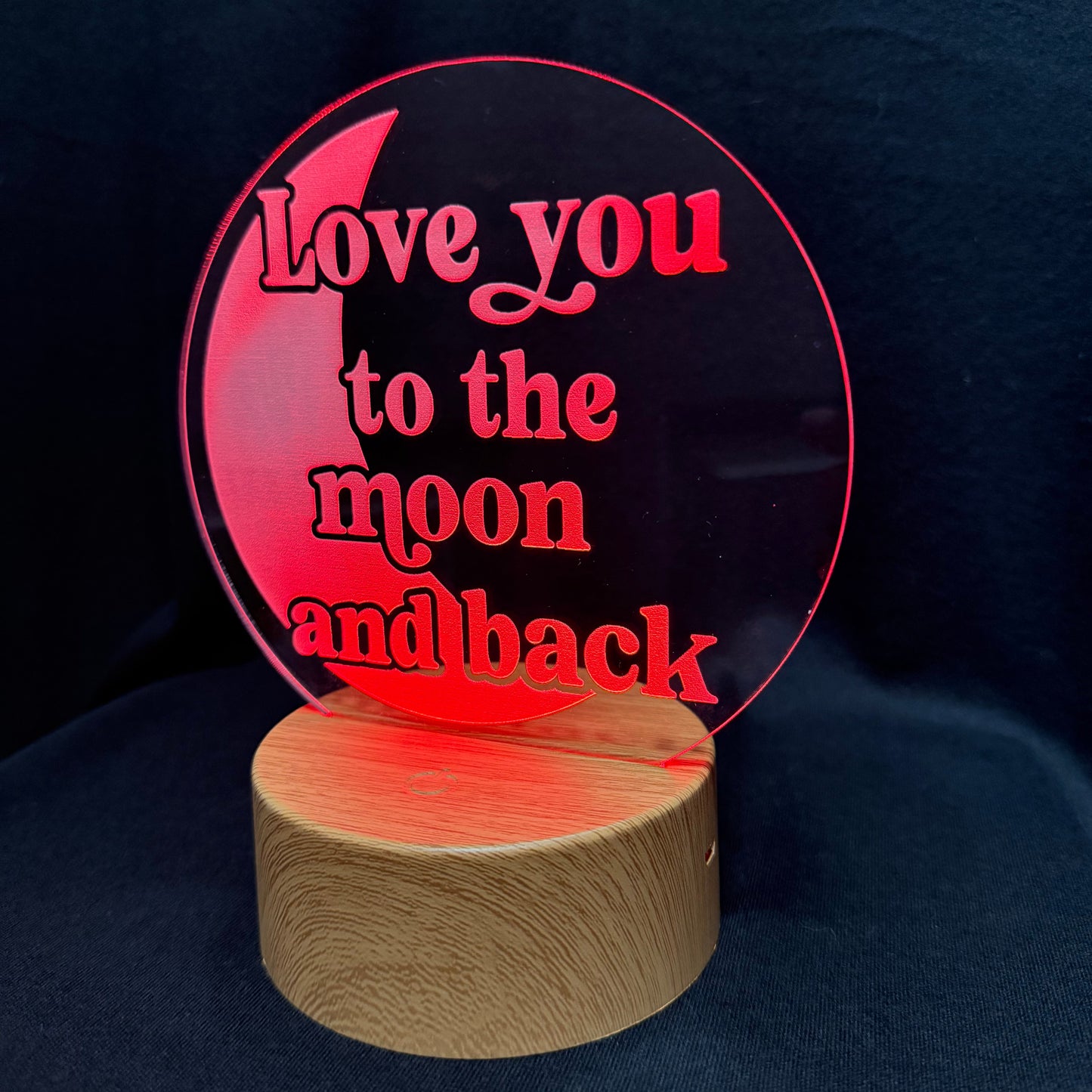 A circular acrylic LED night light with the phrase 'Love you to the moon and back' in red lettering, mounted on a cylindrical wooden base and photographed against a dark fabric background.