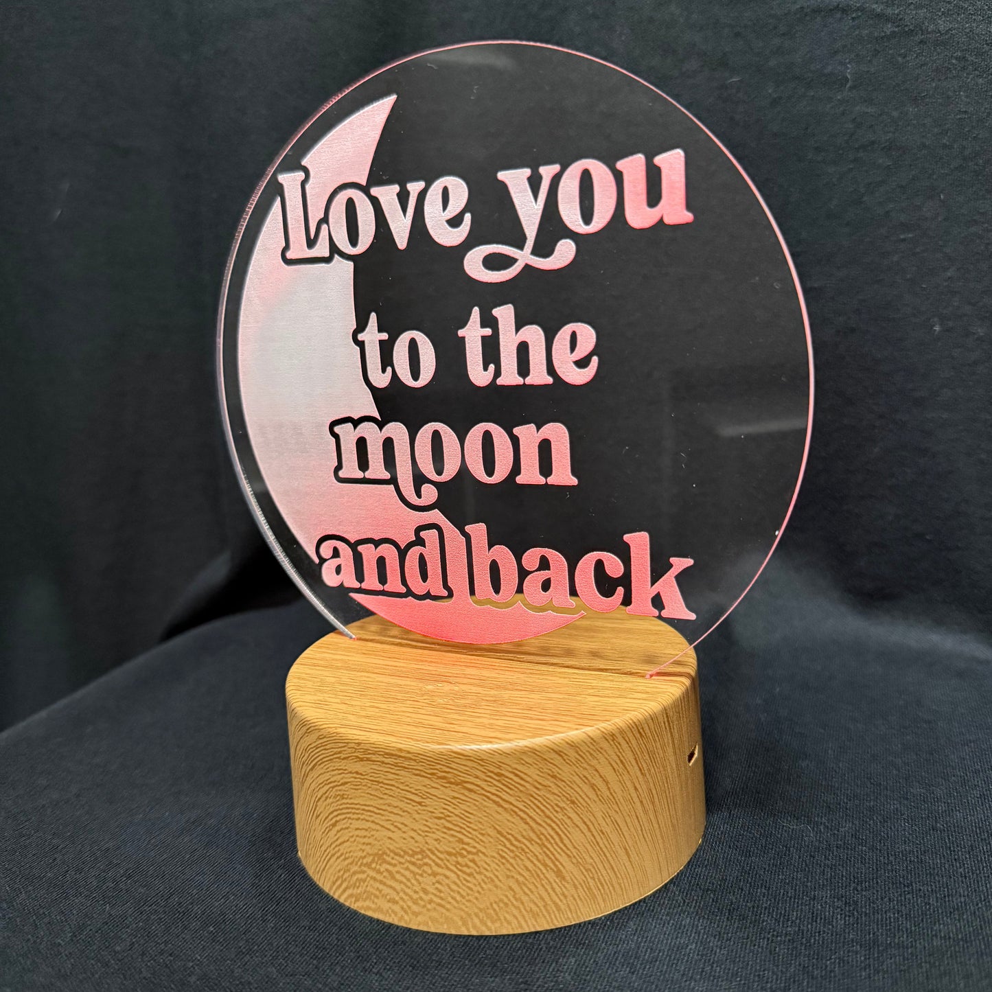 A circular clear acrylic plaque with pink lettering that reads 'Love you to the moon and back,' mounted on a round light wood base against a dark fabric background.