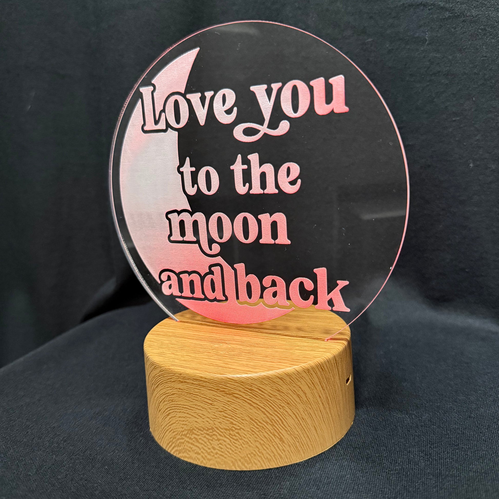 A circular clear acrylic plaque with pink lettering that reads 'Love you to the moon and back,' mounted on a round light wood base against a dark fabric background.