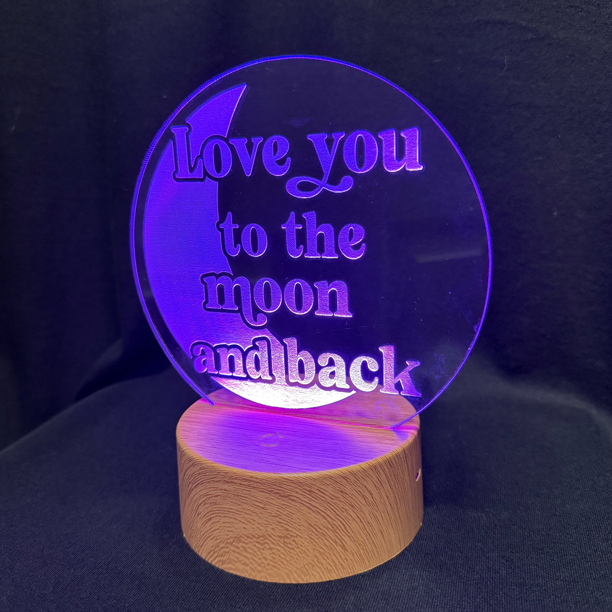 A circular acrylic LED night light lamp with the engraved message 'Love you to the moon and back' glowing purple, resting on a cylindrical wooden base against a dark fabric background.