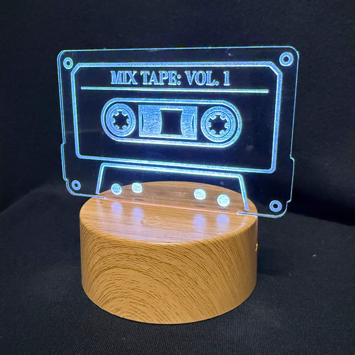 A cassette tape-shaped LED light on a cylindrical wooden base, displaying 'MIX TAPE: VOL. 1' in glowing blue text against a black background.