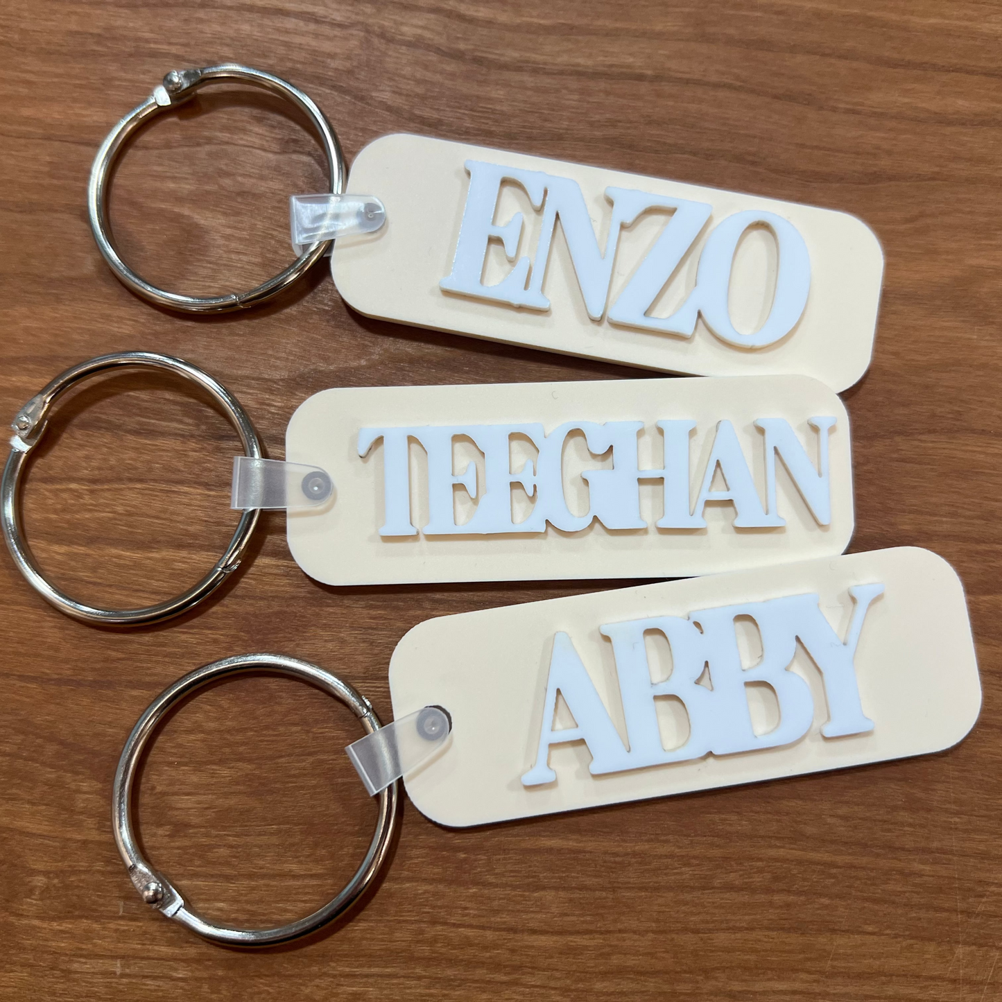 Three beige keychains with cut-out letters spelling 'Abby', 'Teeghan', and 'Enzo' on a wooden surface.