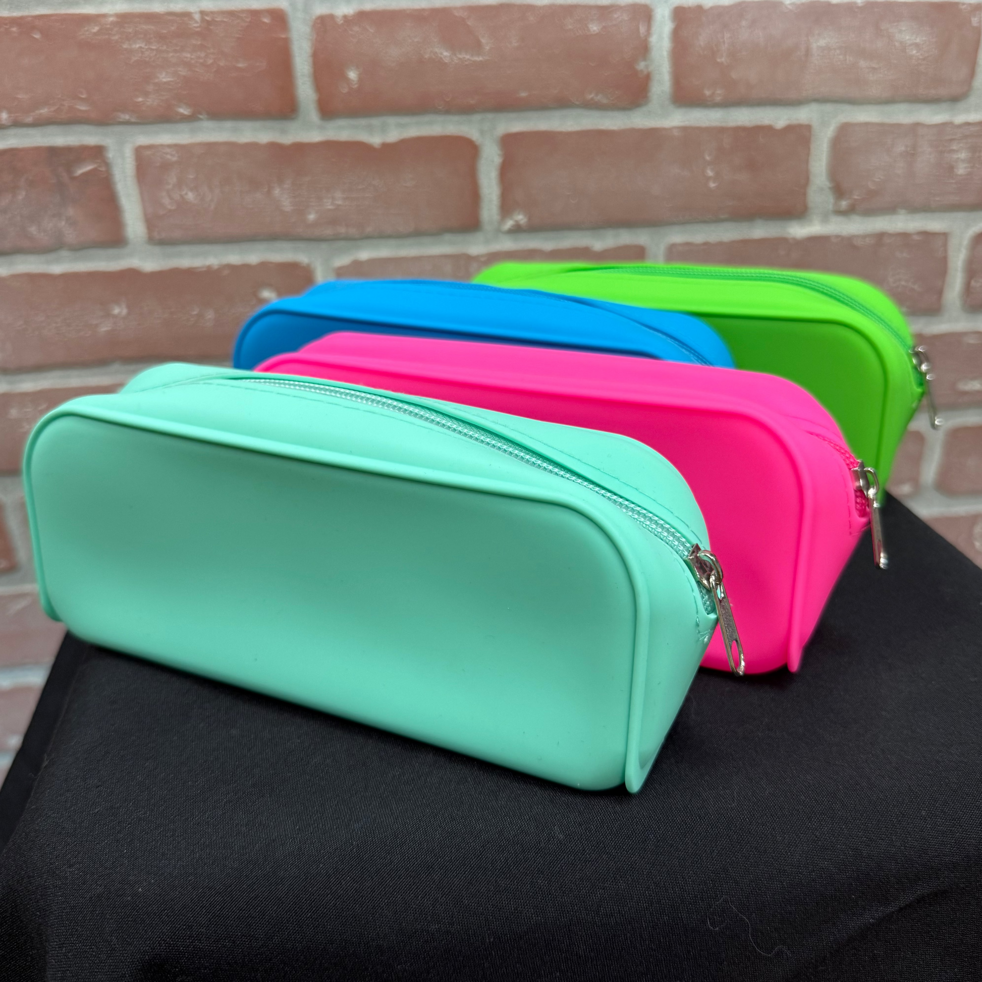 Four zippered pencil cases in mint green, pink, blue, and lime green, arranged in a row on a black cloth surface against a red brick wall background.