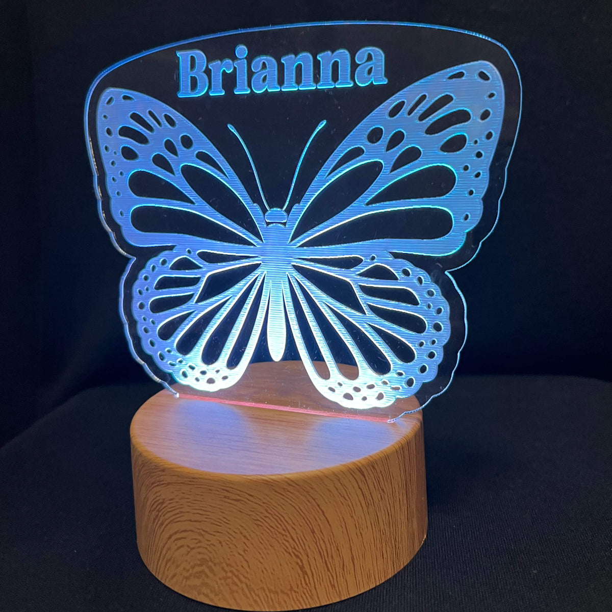 A personalized LED night light featuring a glowing blue butterfly silhouette with the name 'Brianna' cut out at the top, mounted on a cylindrical wooden base against a black background.