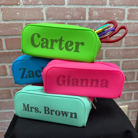 Four personalized zippered pencil cases stacked on a black cloth surface against a brick wall background. The cases are lime green, blue, pink, and mint green, each printed with a name (Carter, Zac, Gianna, Mrs.