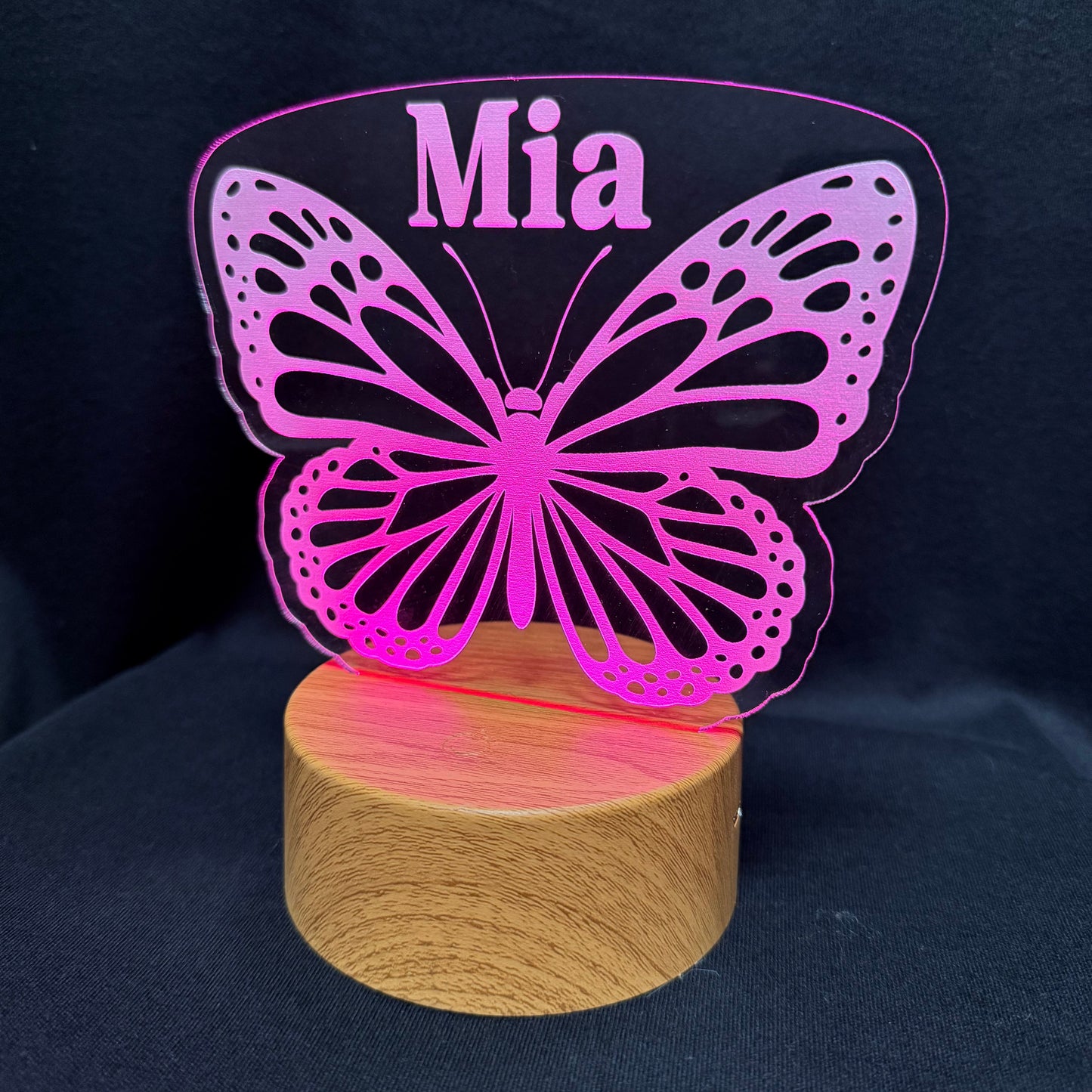 A personalized LED night light featuring a pink butterfly silhouette with the name 'Mia' printed at the top, mounted on a round wooden base against a dark background.
