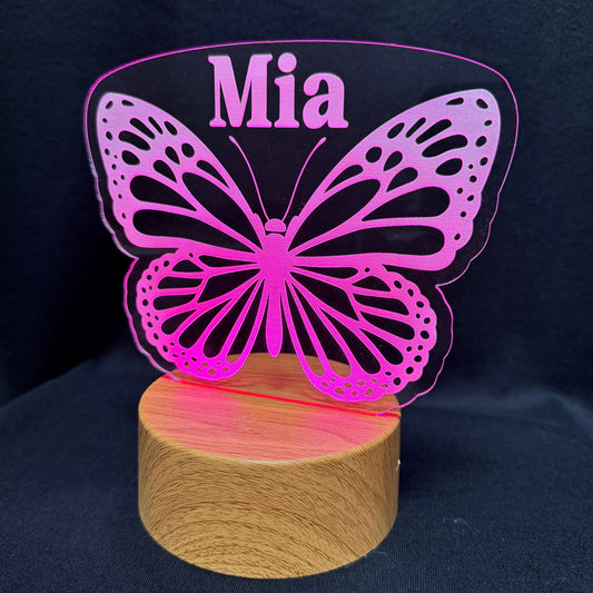 A personalized LED night light featuring a pink butterfly silhouette with the name 'Mia' printed on the top wing section, mounted on a round wooden base against a dark fabric background.