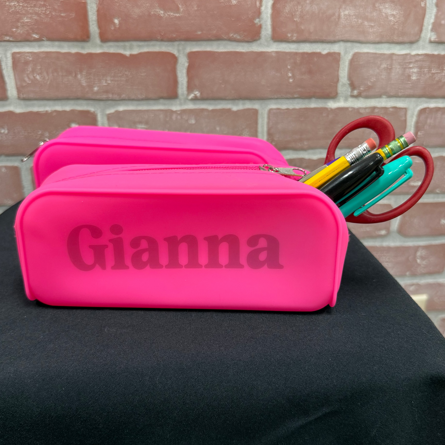 A bright pink personalized pencil case with the name 'Gianna' printed on the front, sitting on a black cloth-covered surface against a red brick wall background. Pens and scissors are tucked into the top.