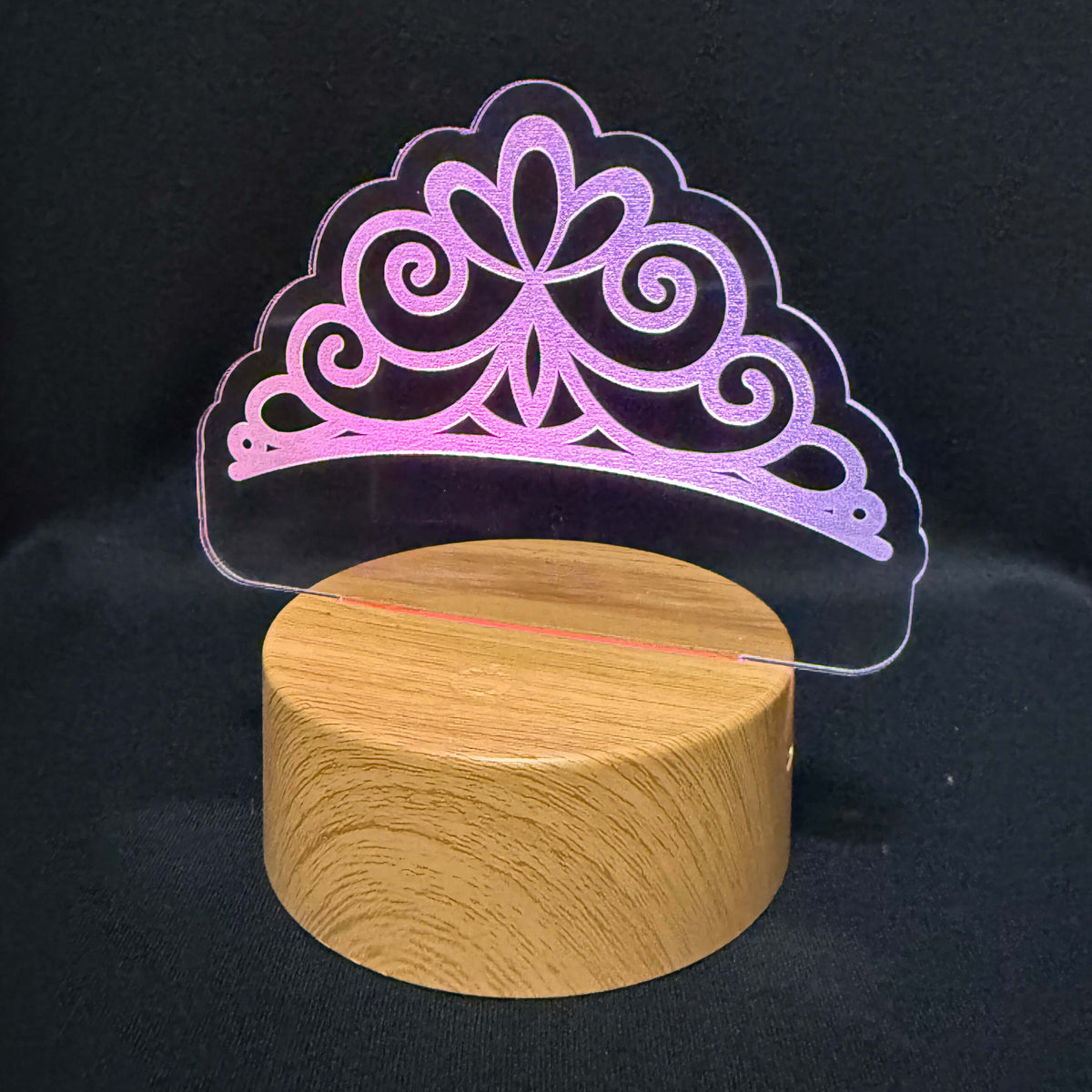 A clear acrylic tiara-shaped lamp with a pink-to-purple illuminated gradient, displayed on a round light wood base against a black fabric background.
