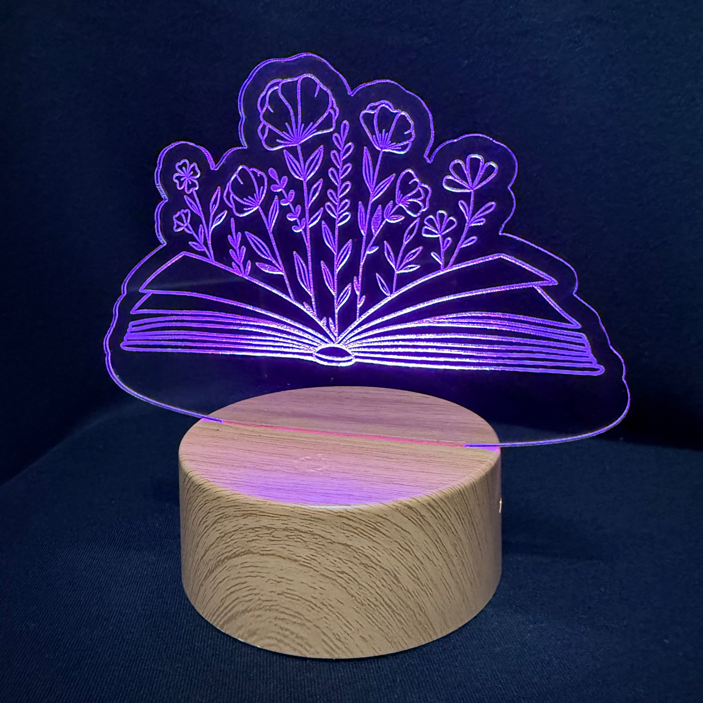 A wooden cylindrical night light base with a clear acrylic plate above it, illuminated by a purple LED line design of an open book with flowers, set against a dark navy background.