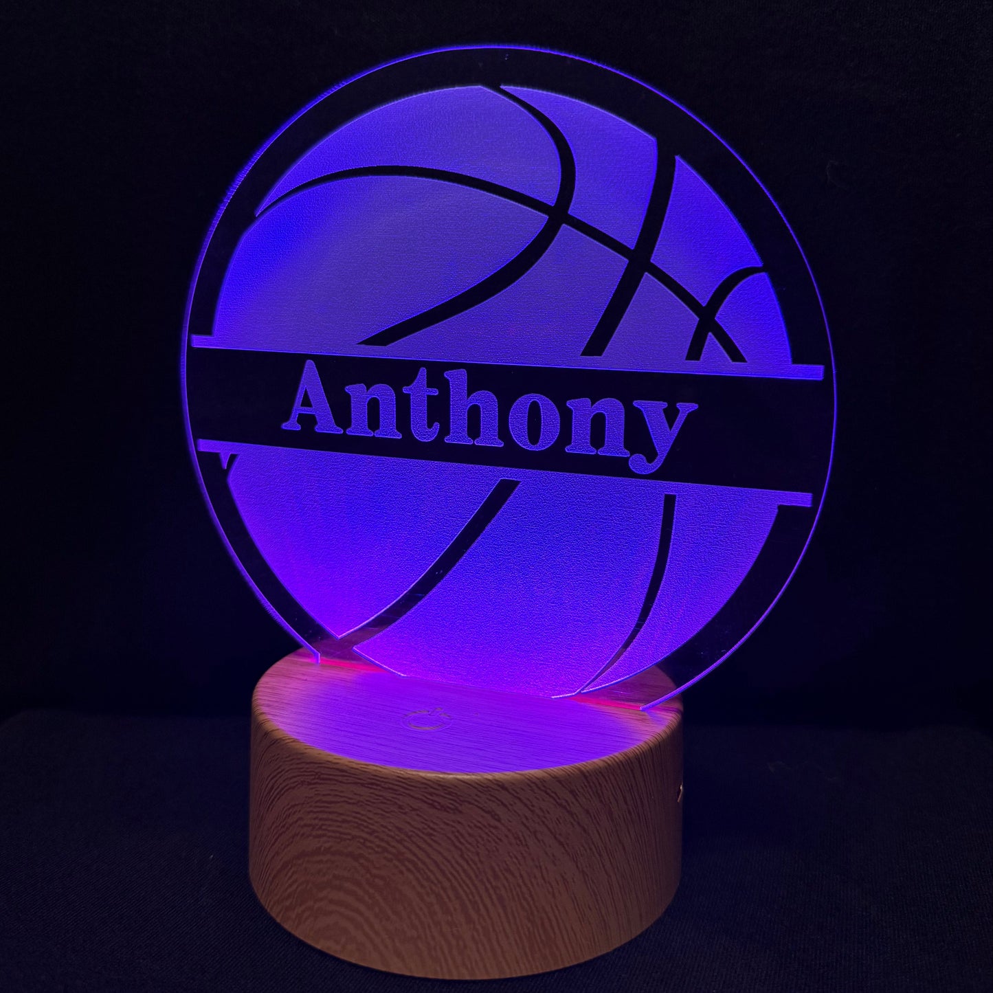 A personalized LED acrylic lamp shaped like a basketball with the name 'Anthony' illuminated in the center, resting on a cylindrical wooden base against a dark background.