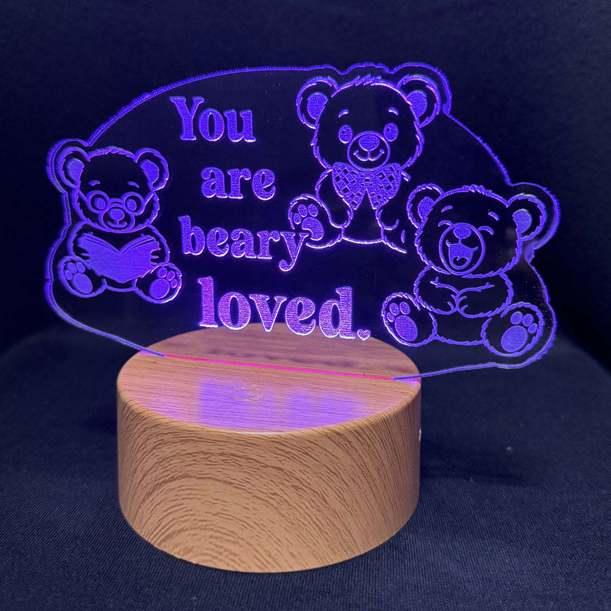 A wooden cylindrical night light base with a clear acrylic plaque above it, illuminated in purple-blue light. The plaque features three teddy bear outlines and the engraved text 'You are beary loved.