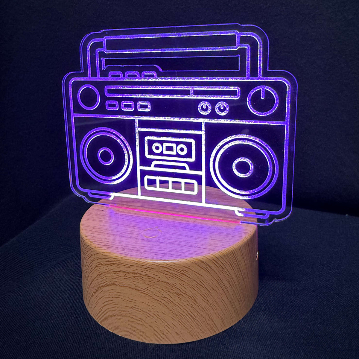 A 3D laser-cut optical illusion lamp shaped like a vintage boombox, glowing with purple light on a cylindrical wooden base against a dark background.