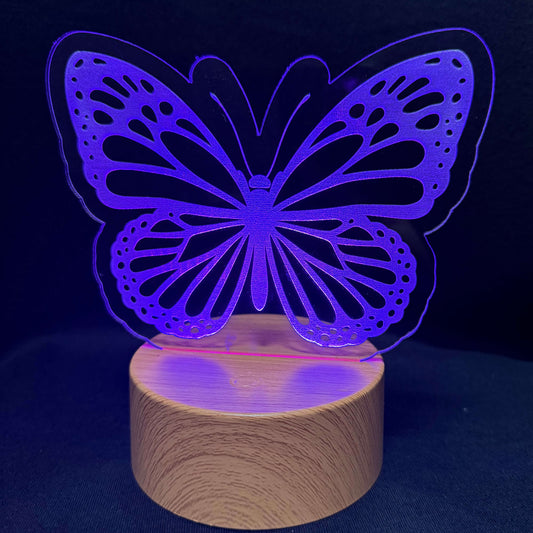 A butterfly-shaped LED night light with a glowing purple laser-cut acrylic top and a cylindrical wooden base, set against a dark blue background.