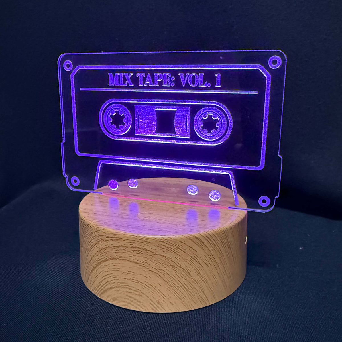 A cassette tape-shaped acrylic light on a cylindrical wooden base, glowing with a purple light against a dark background. The acrylic features the text 'MIX TAPE: VOL.