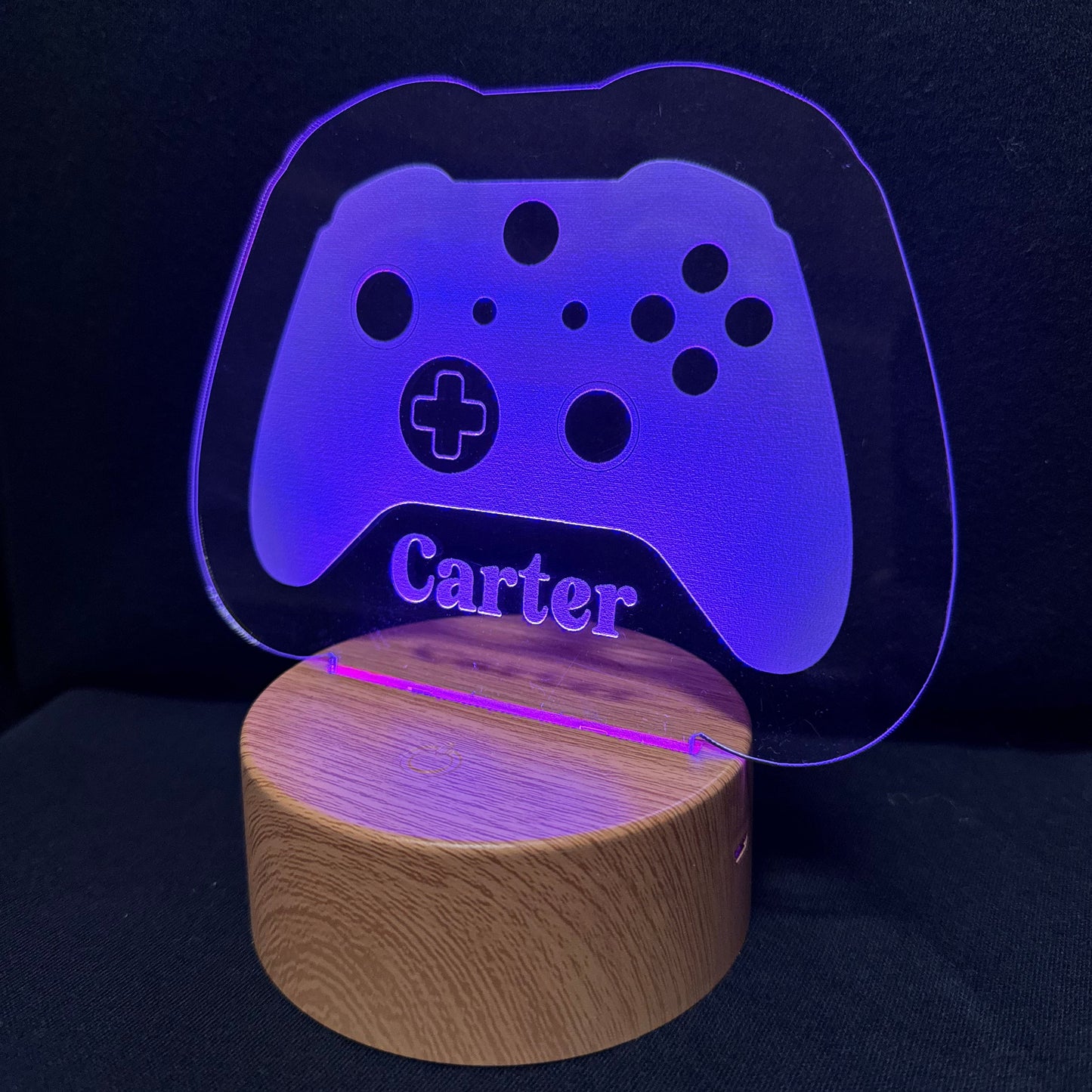 A personalized gaming-themed lamp featuring a clear acrylic panel shaped like a game controller with the name 'Carter' printed on it, illuminated in blue light, resting on a round wooden base against a dark background.