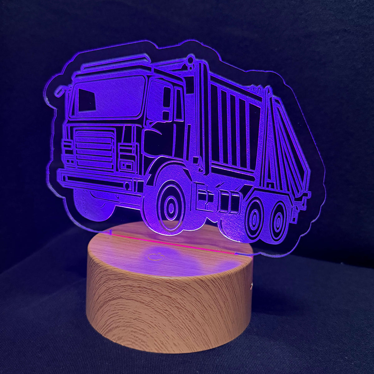 An LED acrylic night light shaped like a garbage truck, glowing with a purple light on a round wooden base against a dark background.