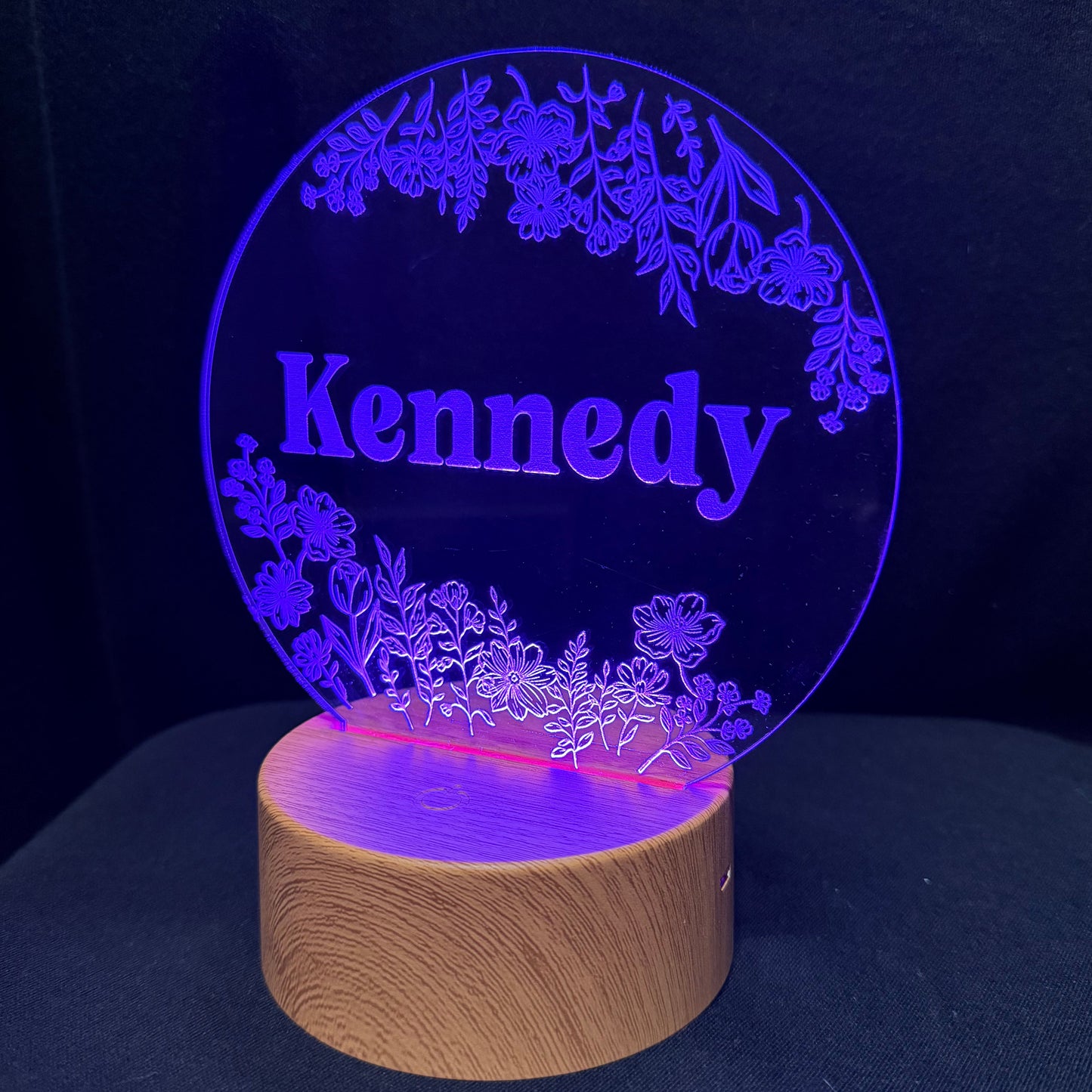 A personalized LED acrylic night light featuring the name 'Kennedy' in purple text, surrounded by floral designs, mounted on a round wooden base and illuminated against a dark background.
