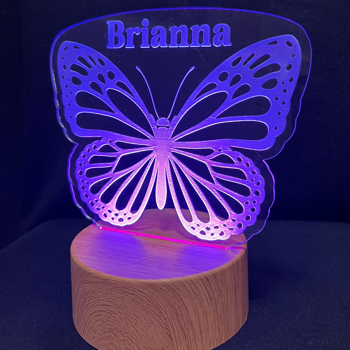 A personalized LED acrylic night light shaped like a butterfly with the name 'Brianna' engraved at the top, mounted on a round wooden base and glowing with purple and blue light against a dark background.