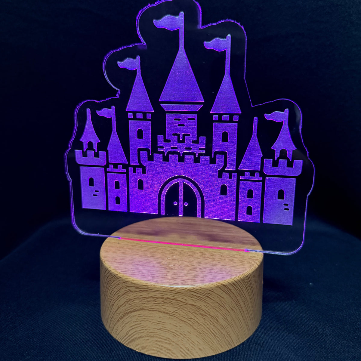 A castle-shaped acrylic LED night light glowing with purple and pink light, mounted on a round wooden base against a dark background.