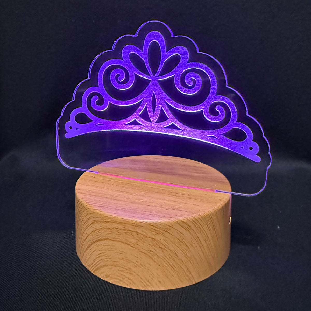 A 3D optical illusion night light featuring a glowing purple tiara design projected above a round wooden base, set against a dark black background.