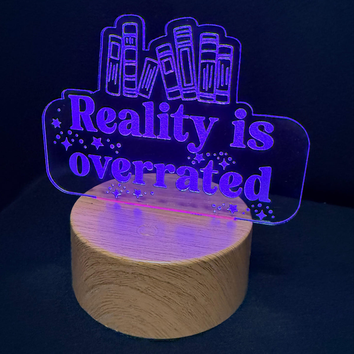 A wooden cylindrical lamp base with a glowing acrylic sign that reads 'Reality is overrated' in purple light, set against a dark navy background.