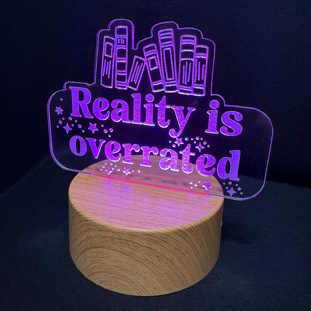 An LED acrylic night light with the text 'Reality is overrated' in purple, featuring a wooden cylindrical base and a laser-cut skyline design on a dark background.