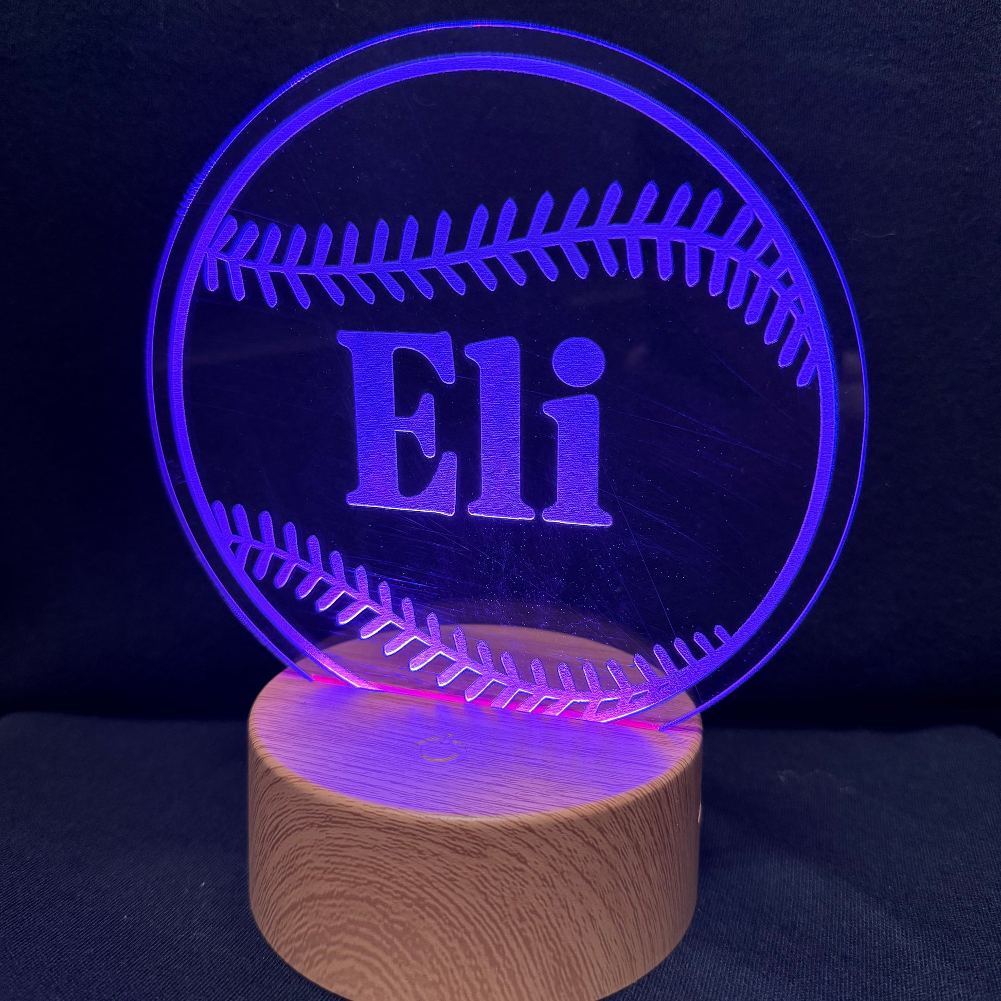 A personalized baseball-shaped LED night light on a wooden base, featuring the name 'Eli' illuminated in purple against a dark background.