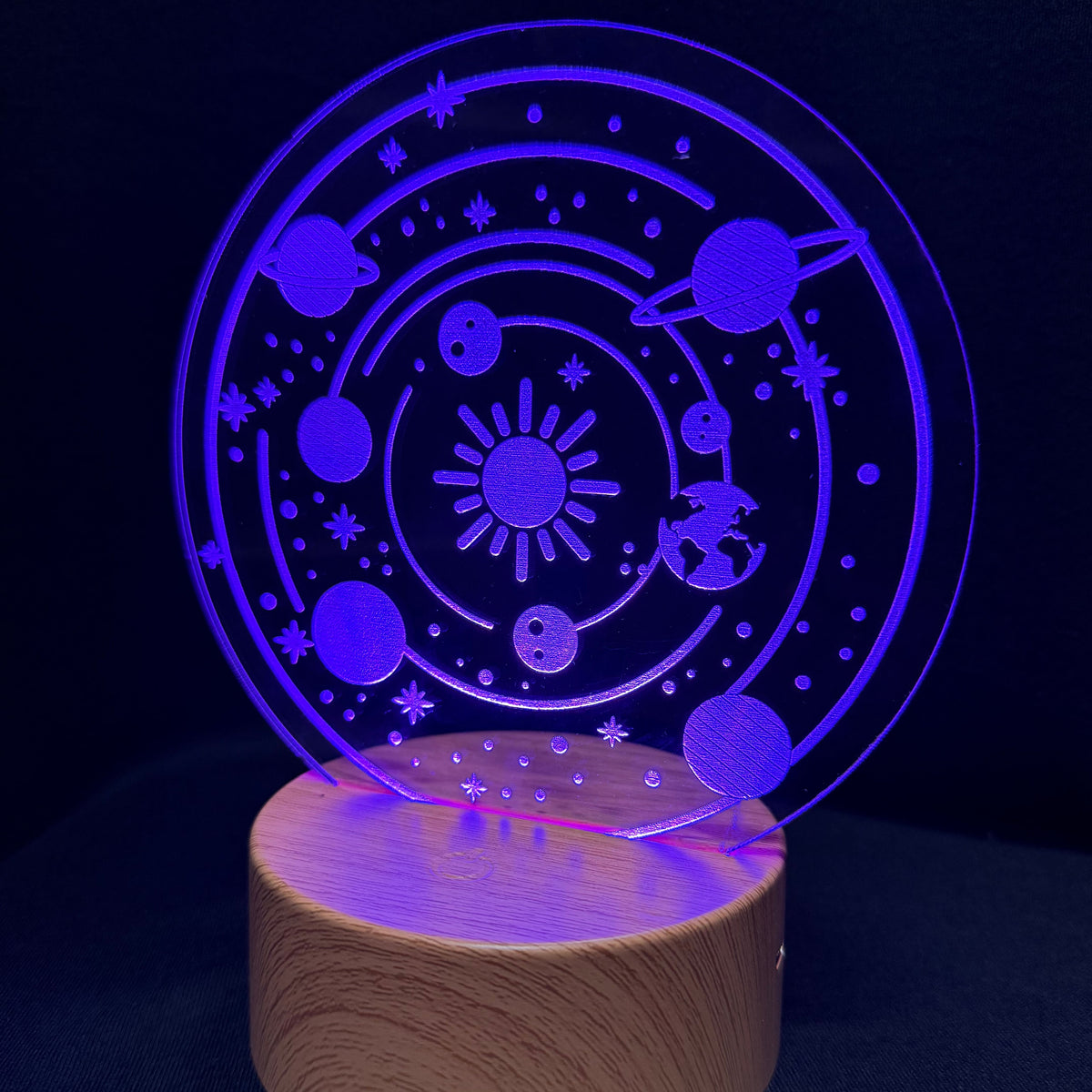 A 3D optical illusion night light featuring a solar system design with planets, stars, and a sun, illuminated in a vibrant purple-blue light on a round wooden base against a dark background.