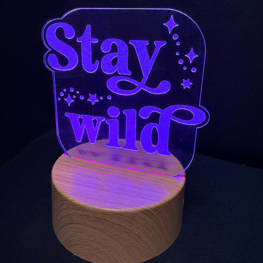 A 3D acrylic LED night light with the phrase 'Stay wild' in glowing purple lettering, mounted on a cylindrical wooden base and photographed against a dark background.