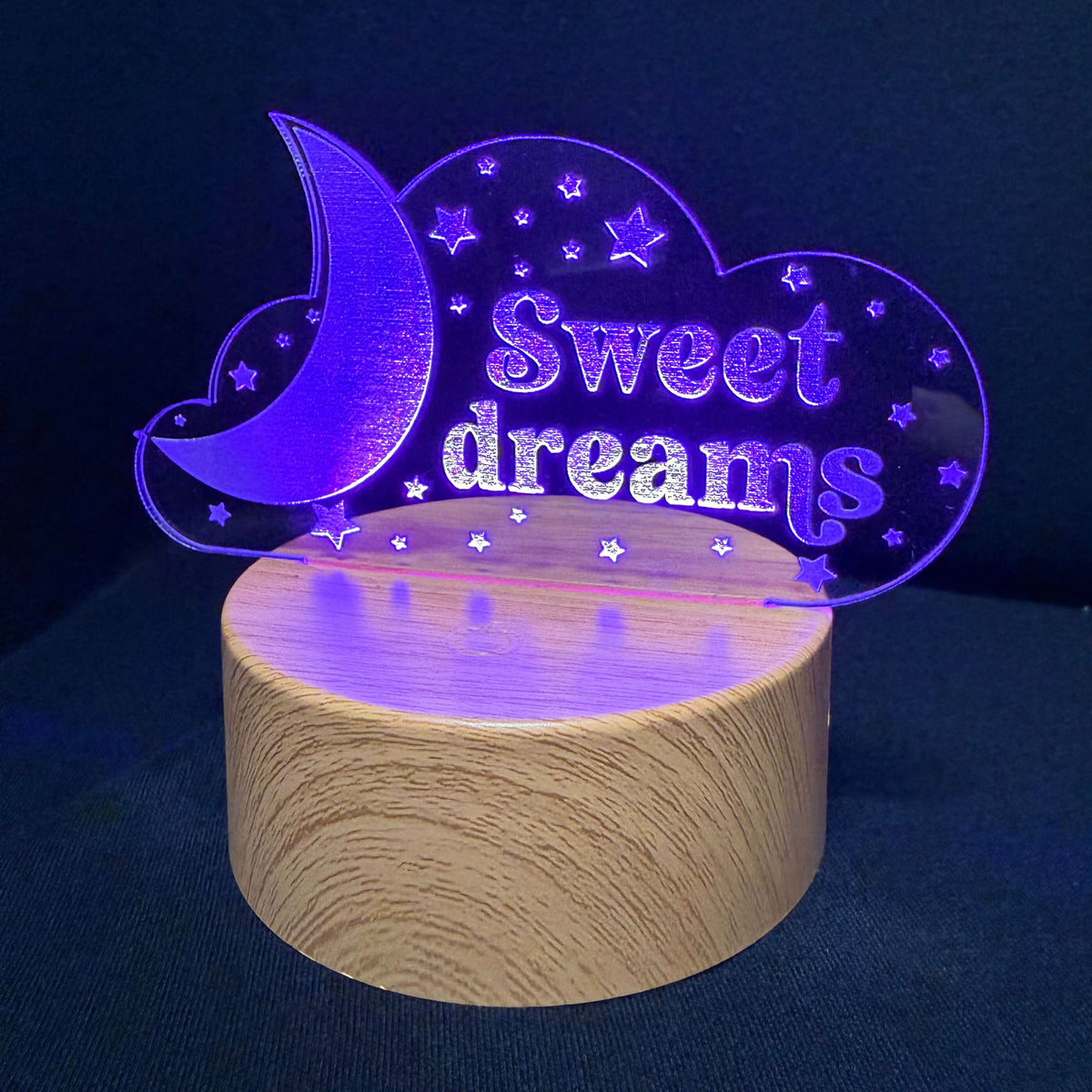 A 3D laser-engraved acrylic night light shaped like a crescent moon and cloud with the words 'Sweet dreams' and small stars, illuminated in purple light and mounted on a cylindrical wooden base against a dark blue background.