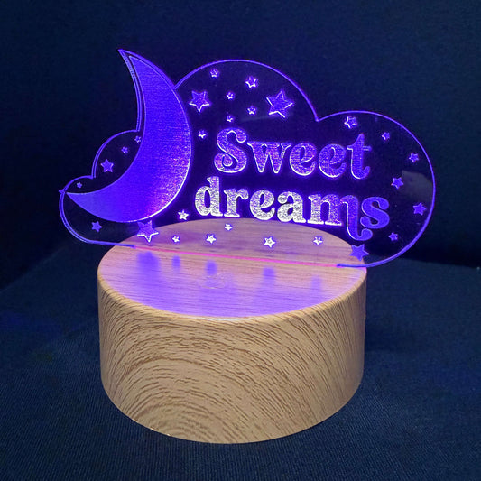 A 3D laser-engraved acrylic night light shaped like a crescent moon and cloud with the words 'Sweet dreams' and small stars, illuminated in purple light and mounted on a cylindrical wooden base against a dark blue background.