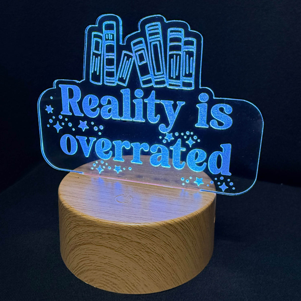 A novelty LED night light with a wooden base and a clear acrylic sign that reads 'Reality is overrated' in glowing blue text, set against a dark background.