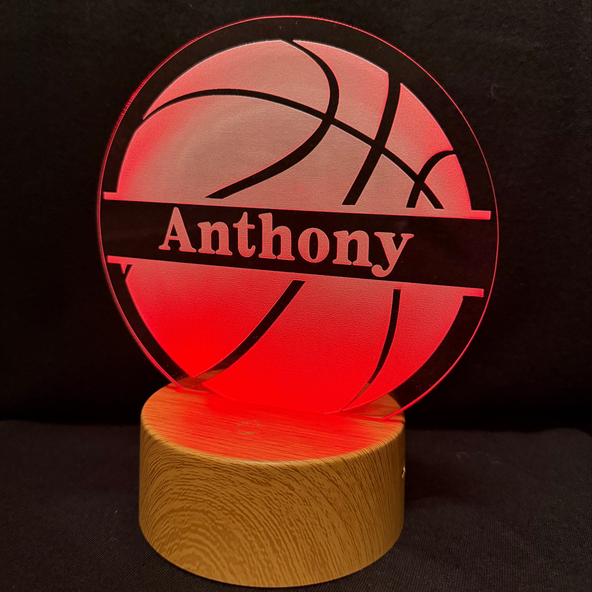 A personalized LED acrylic night light shaped like a basketball, glowing with a red light and featuring the name 'Anthony' in white text on a black band. The light sits on a cylindrical wooden base against a dark background.