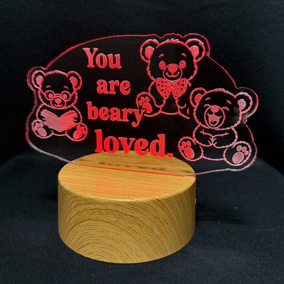 A personalized acrylic LED night light featuring three teddy bear illustrations and the text 'You are beary loved.' in red, mounted on a cylindrical light-wood base against a black background.