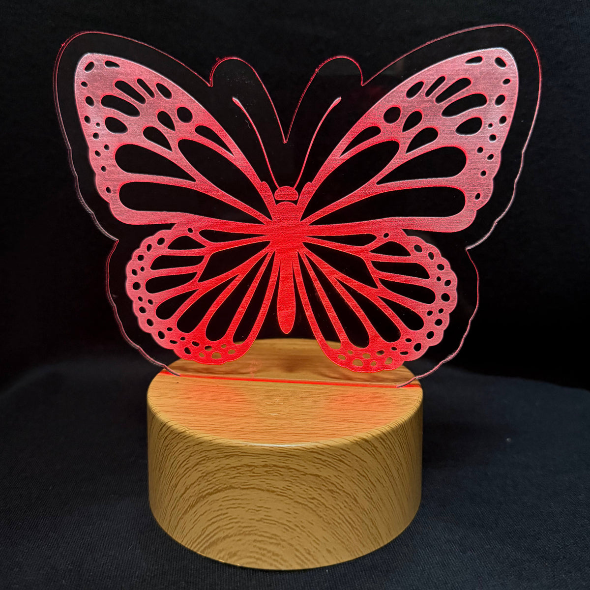 A red laser-cut butterfly acrylic lamp on a round wooden base, set against a dark black background.
