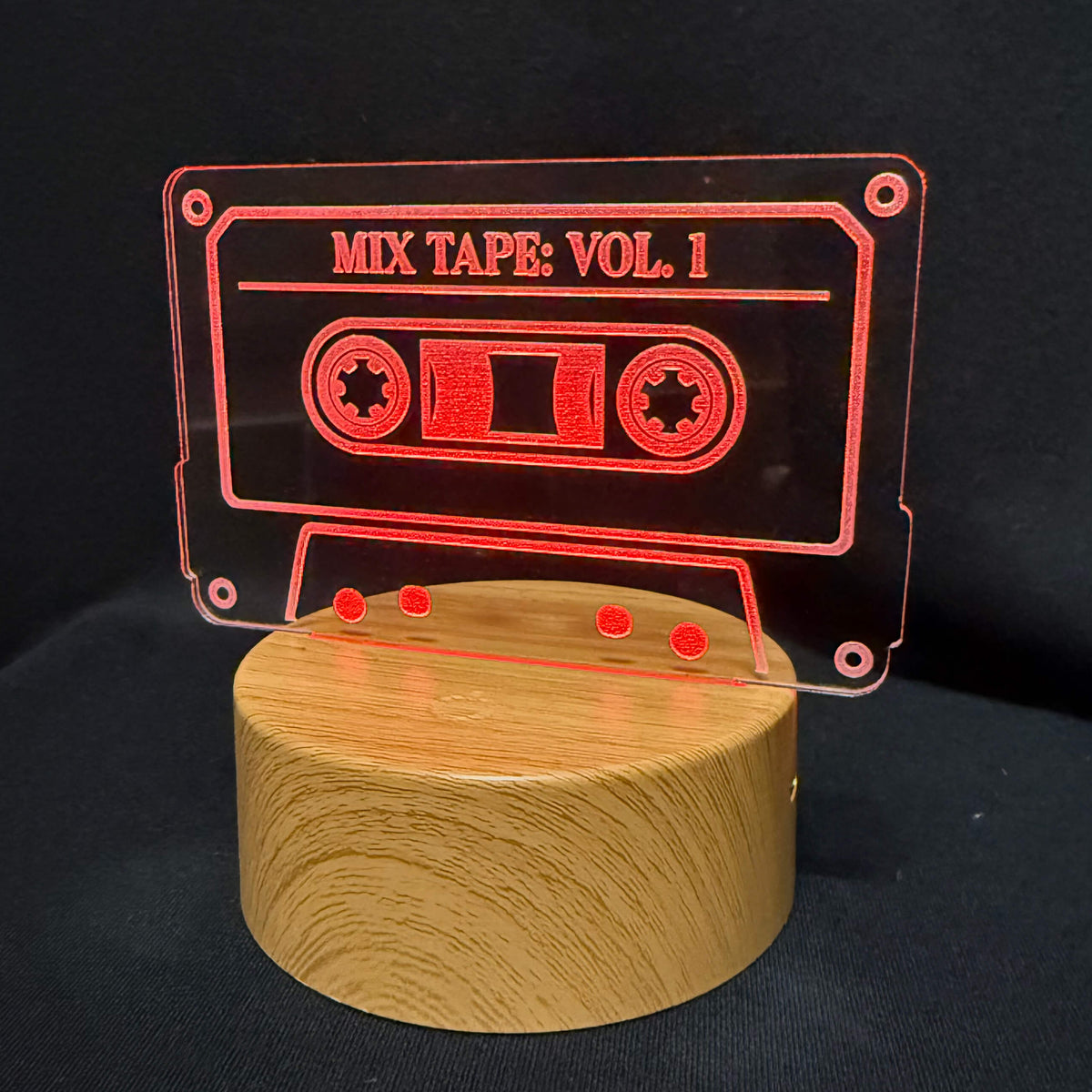 A retro cassette tape-shaped acrylic wall light with 'MIX TAPE: VOL. 1' cut out, mounted on a round wooden base against a black background.