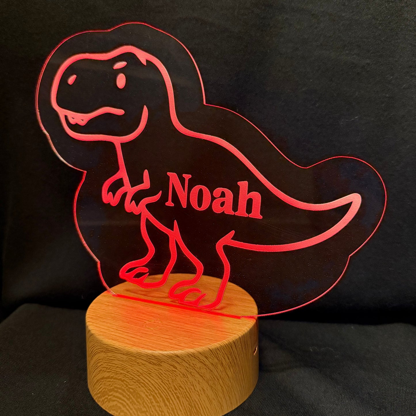 A personalized acrylic T-Rex dino lamp with the name 'Noah' cut out, mounted on a round wooden base and illuminated with a red glow against a black background.