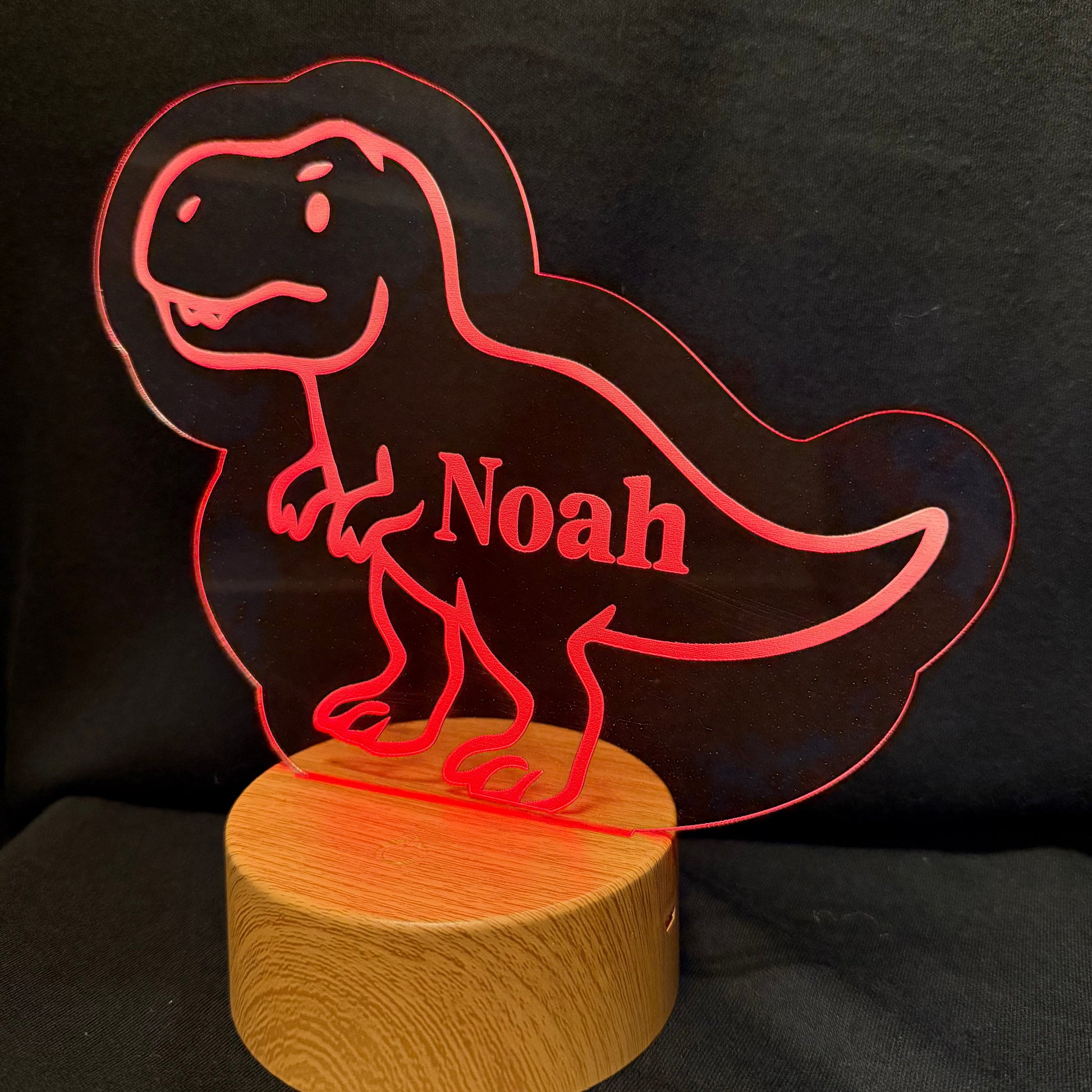 A personalized acrylic T-Rex dino lamp with the name 'Noah' cut out, mounted on a round wooden base and illuminated with a red glow against a black background.
