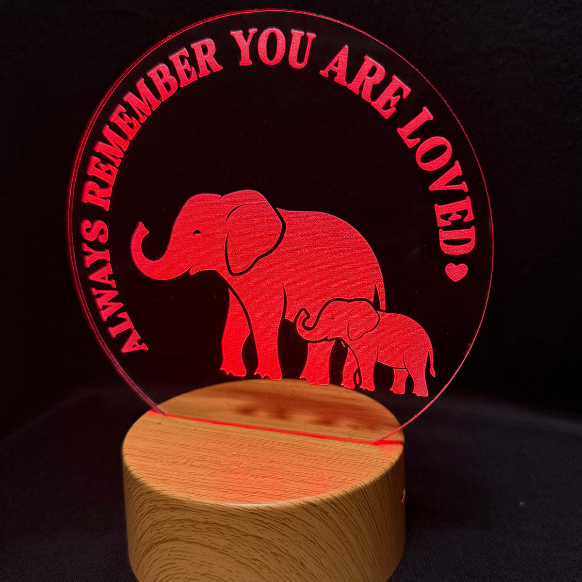 A red acrylic sign featuring a mother and baby elephant silhouette with the text 'ALWAYS REMEMBER YOU ARE LOVED' in red, mounted on a round wooden base against a black background.