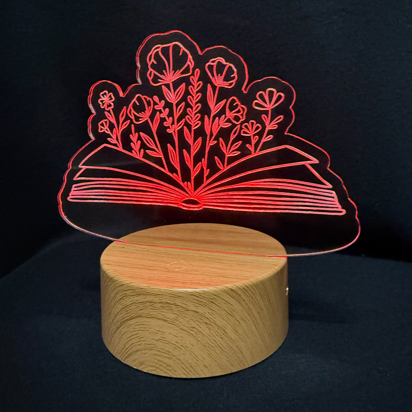 A wooden cylindrical night light base with a clear acrylic plate featuring a red illuminated design of an open book and flowers, set against a dark background.