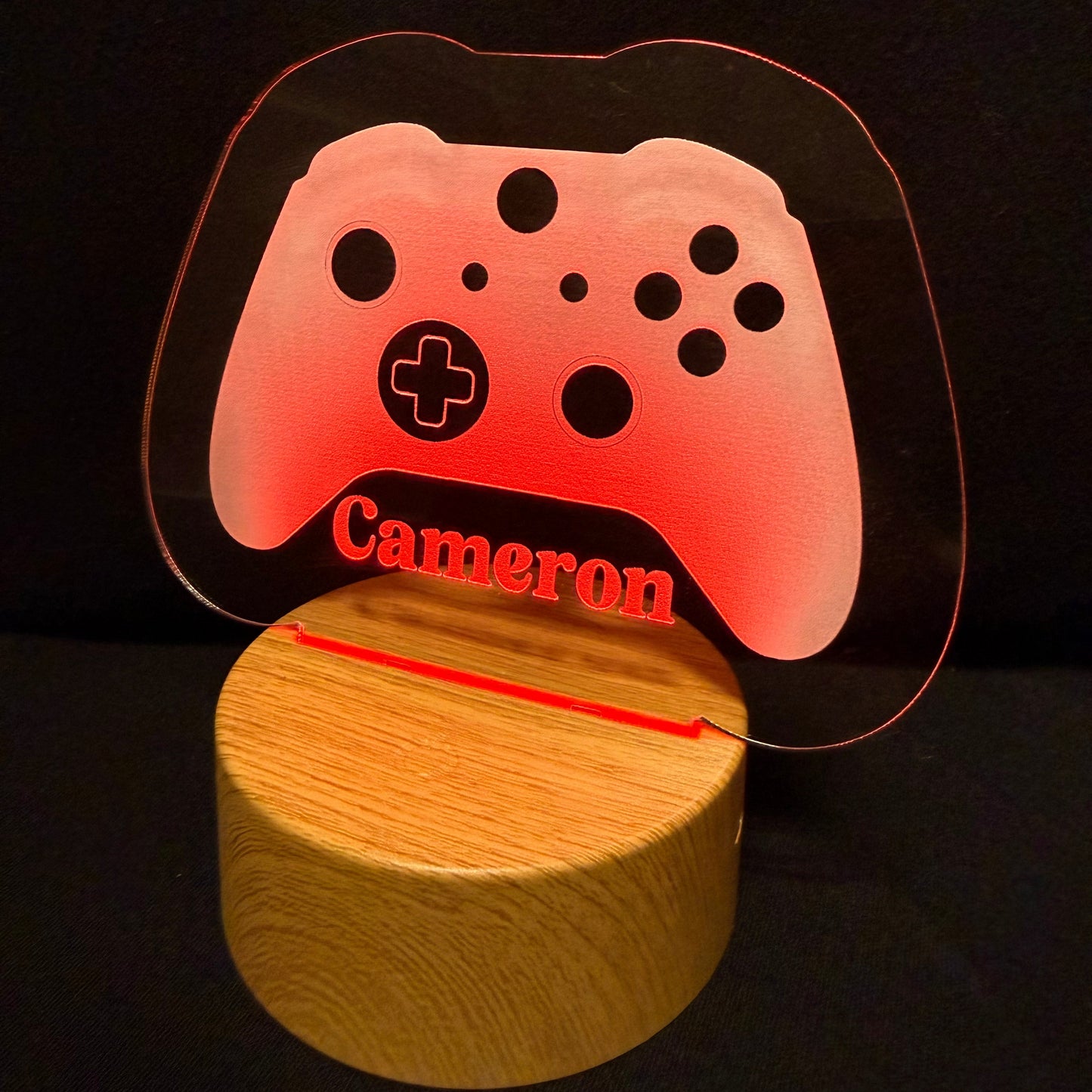 A personalized gaming-themed LED night light featuring a translucent acrylic controller-shaped panel with the name 'Cameron' in red, illuminated with a warm pink glow, mounted on a round wooden base against a black background.