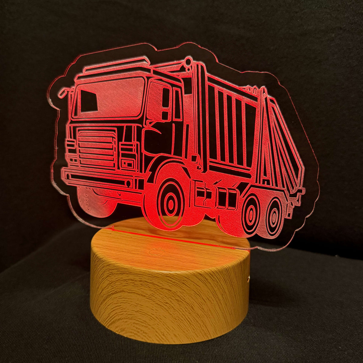 A red laser-cut acrylic sign depicting a garbage truck, mounted on a round wooden base and illuminated against a dark background.