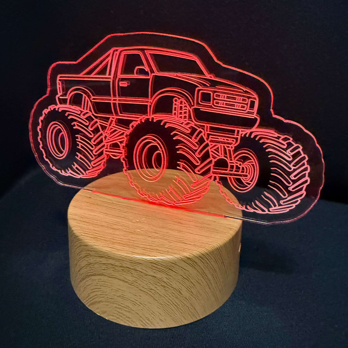 A 3D laser-engraved acrylic light shaped like a monster truck, glowing red, mounted on a round wooden base against a dark background.