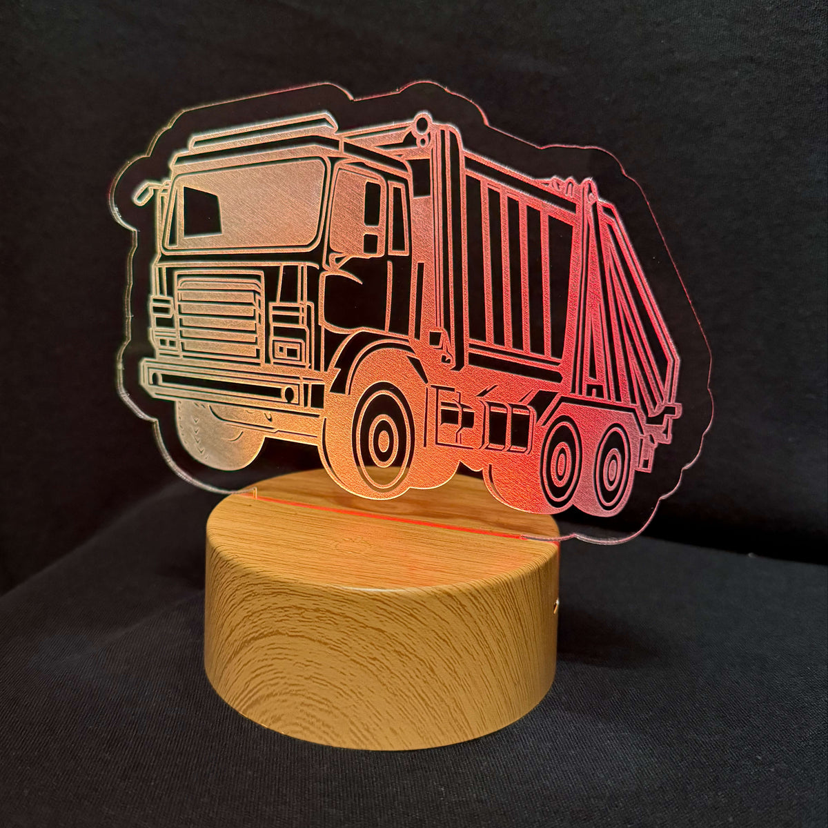 An acrylic laser-cut lamp featuring a garbage truck design, illuminated with a warm pink and orange glow, resting on a round wooden base against a dark background.