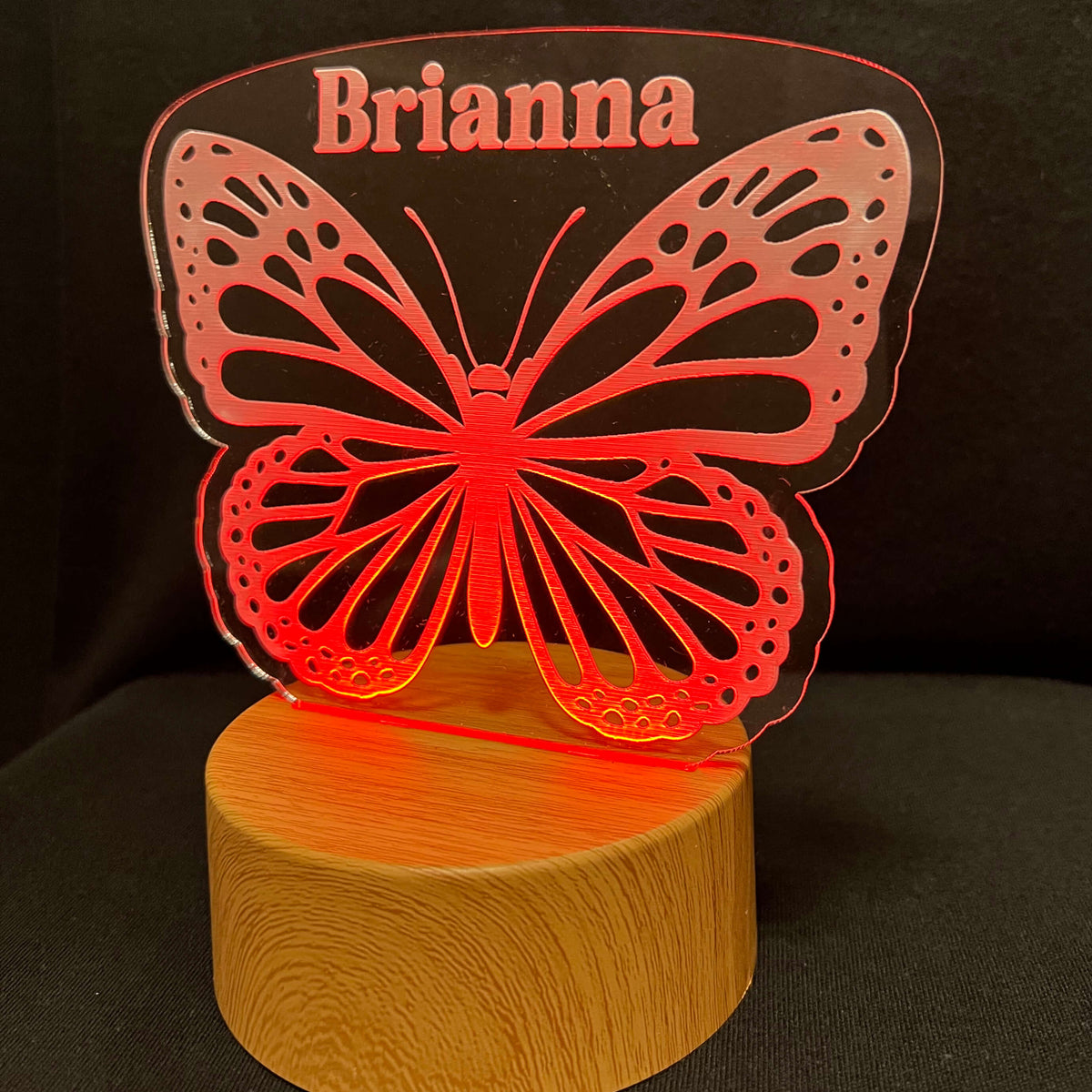 A personalized LED butterfly night light with a wooden base and a clear acrylic butterfly-shaped top featuring the name 'Brianna' in red, shown on a black background.