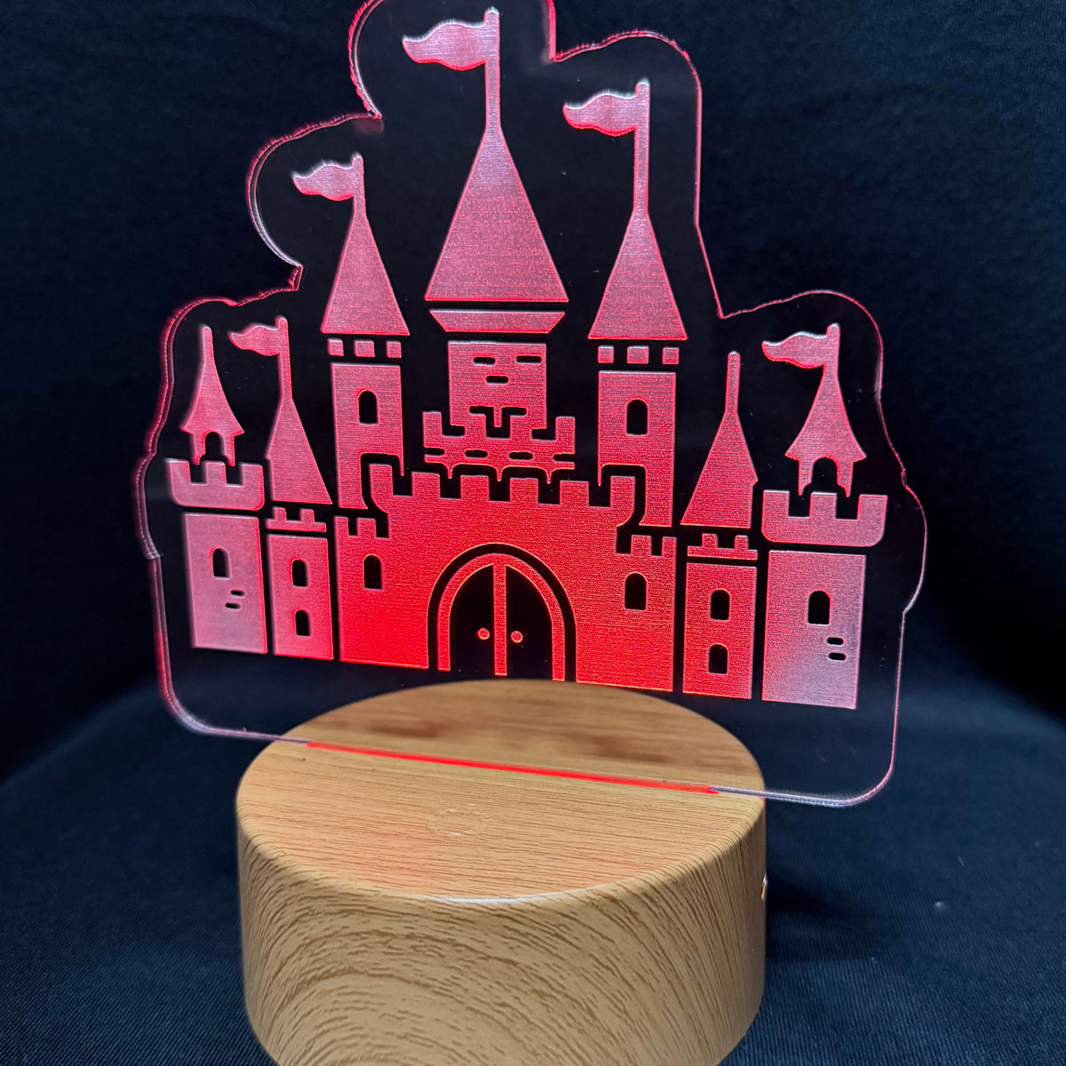A castle-shaped acrylic night light with a wooden base, illuminated with a pink and red glow against a dark blue background.