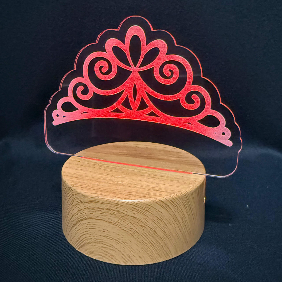 A red laser-cut acrylic tiara-shaped decorative piece displayed on a round wooden base against a dark navy fabric background.